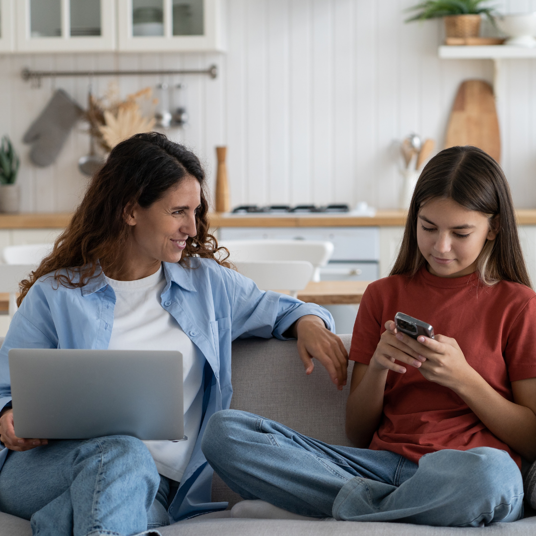 Mother sitting on couch with computer and daughter sitting with cell phone.
