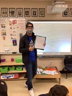 A teacher stands in front of a class holding a small whiteboard